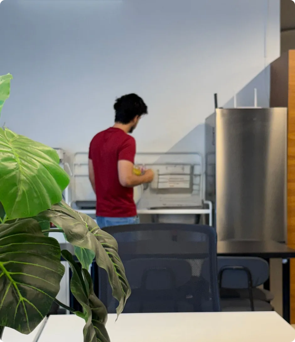 Man in a red shirt standing in front of a stainless steel appliance in a room with plants and chairs.