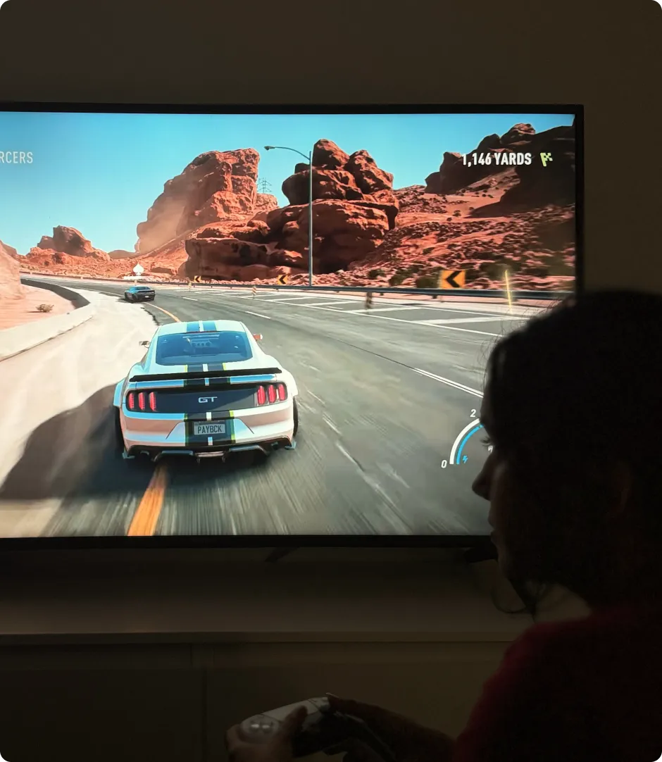Person holding a game controller playing a car racing video game featuring a white GT car with a black and yellow stripe on a desert road.