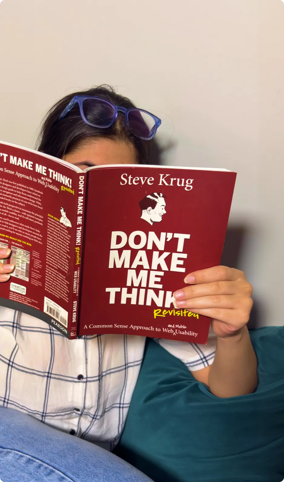 Person wearing a white plaid shirt and blue glasses on head reading the book 'Don't Make Me Think' by Steve Krug.