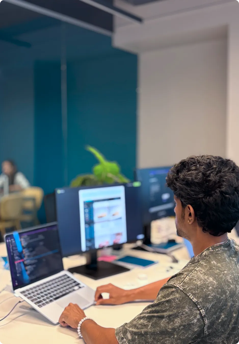 Man working on a laptop and two monitors at a desk in an office with a blue accent wall and blurred background.