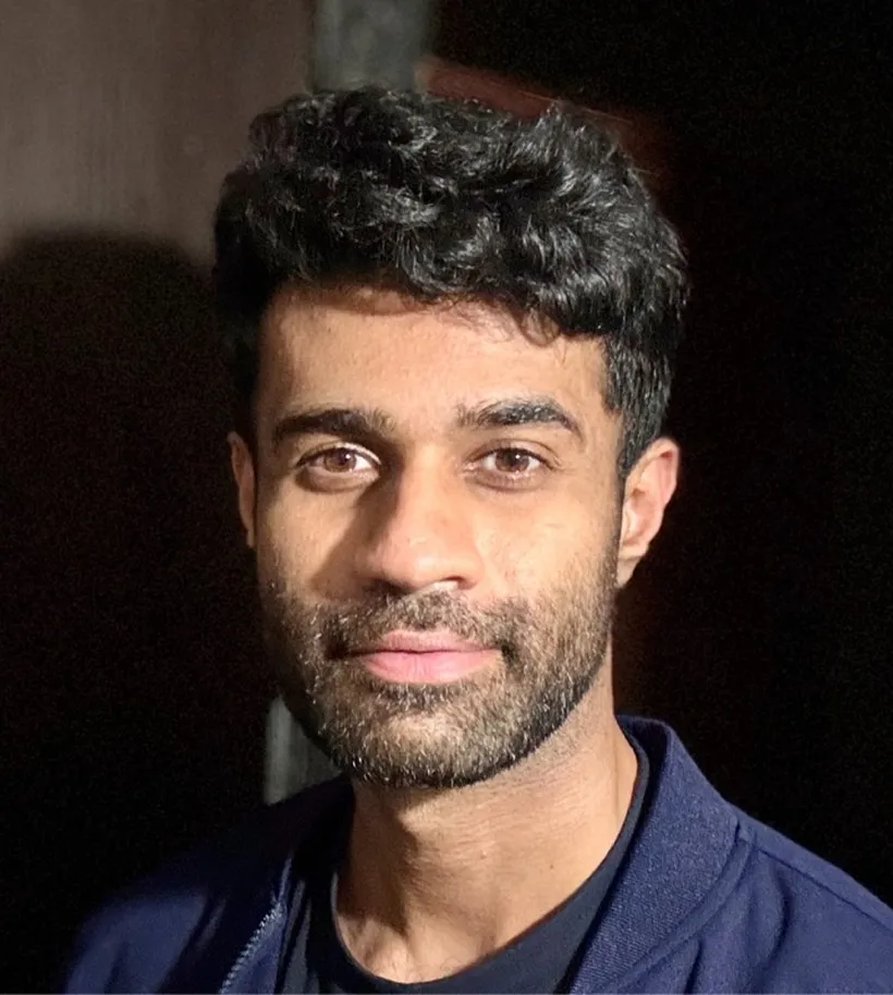 Close-up of a young man with curly dark hair, trimmed beard, and a slight smile against a dark background.