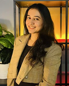Smiling woman with long dark hair wearing a plaid blazer, standing indoors by a plant and warm light.