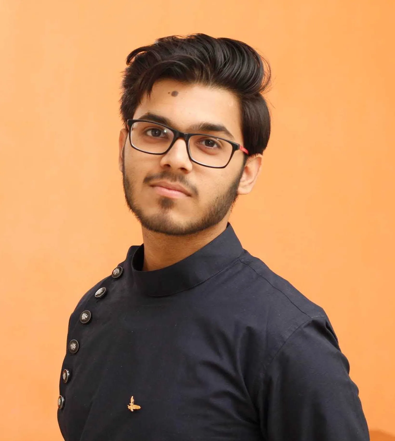 Young man with glasses and neatly styled hair wearing a dark buttoned shirt against an orange background.