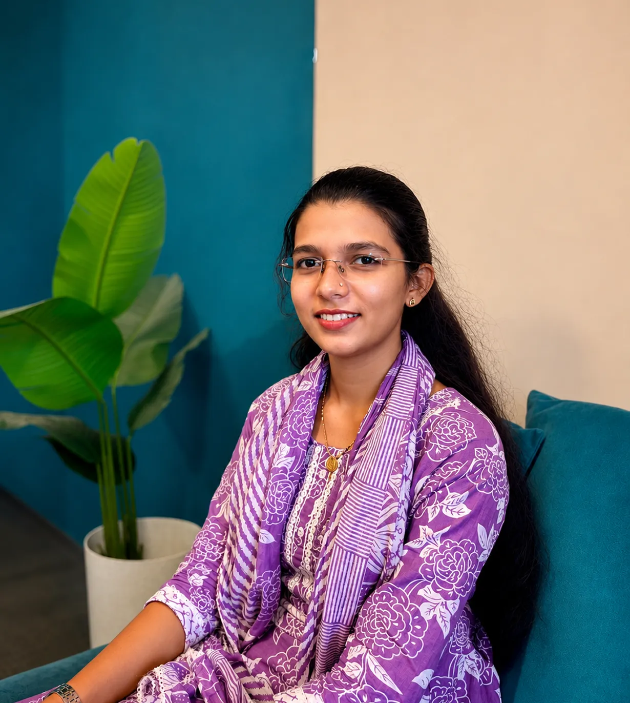 Woman wearing glasses and a purple floral outfit sitting on a teal chair with a green plant nearby.