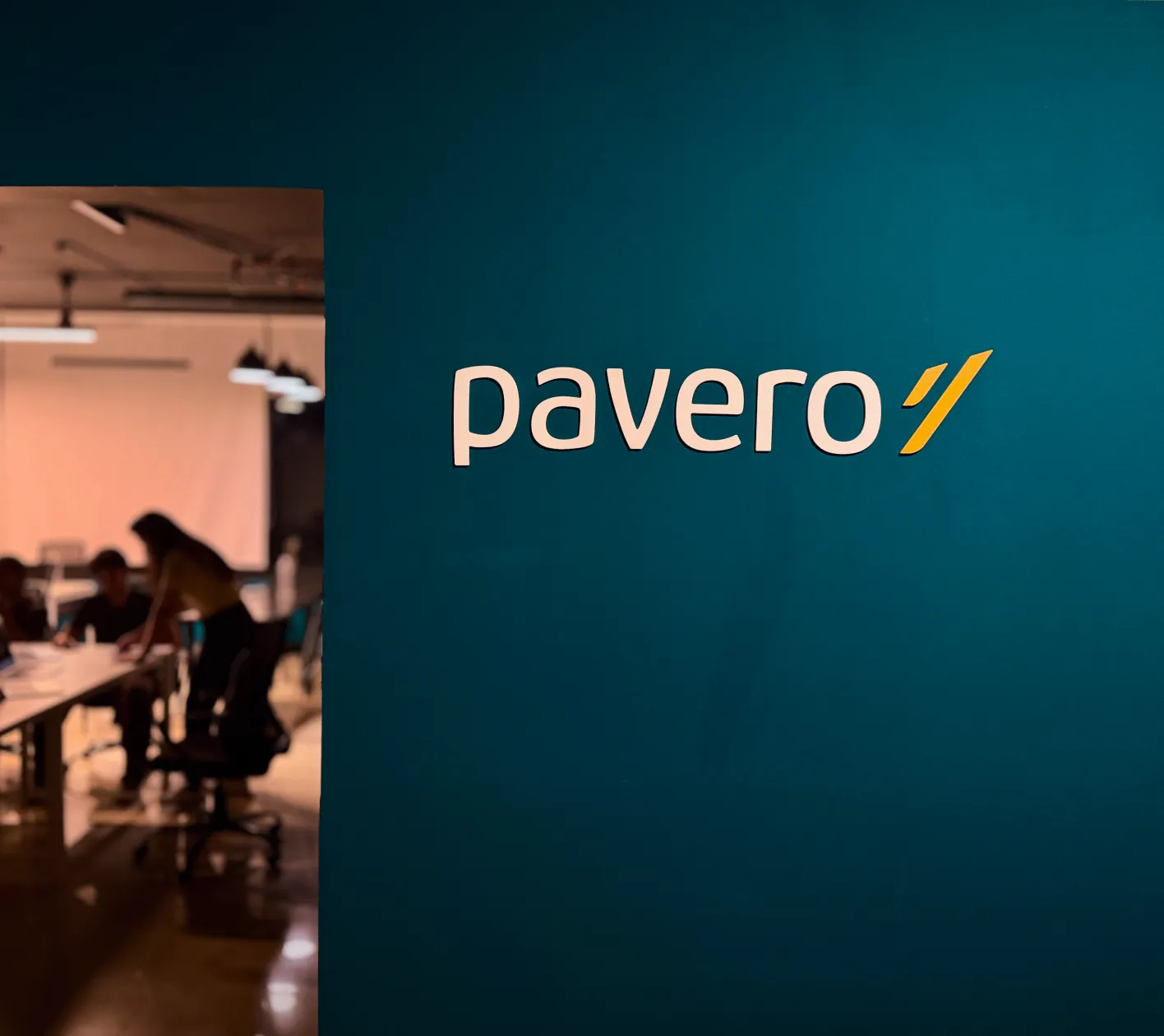 Blue wall with the word 'pavero' and a yellow logo; blurred office scene with people working in background