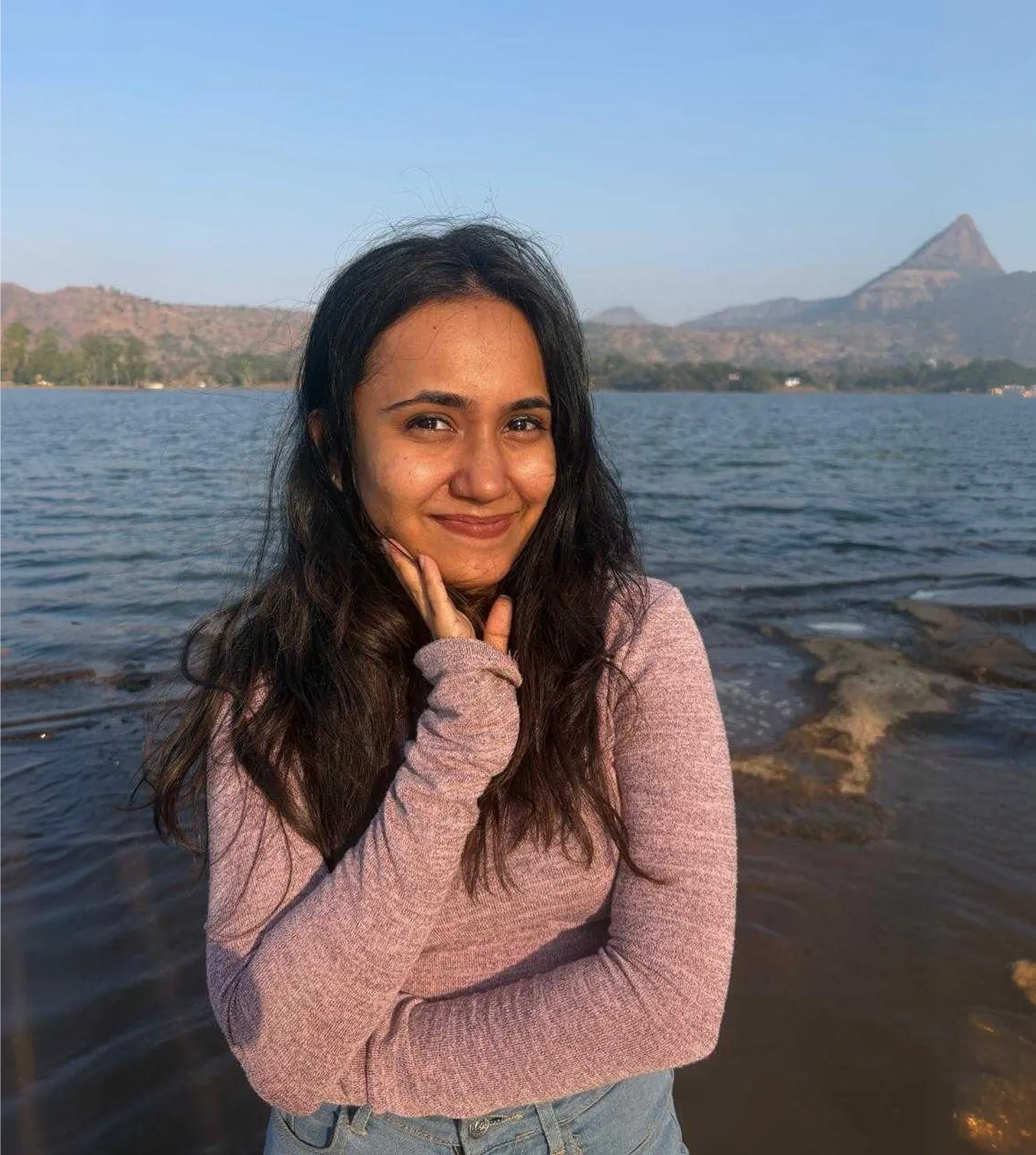 Woman smiling with long hair, wearing a mauve sweater and jeans by a lake with hills in the background.