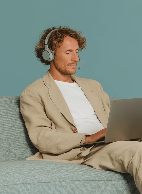 A professionally dressed man using a laptop with headphones while sitting on a sofa, highlighting remote hiring efficiency and modern workplace flexibility.