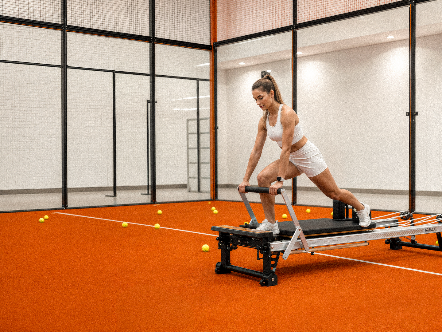 Woman in white sportswear exercises on a Pilates reformer in a room with orange floor and tennis balls.