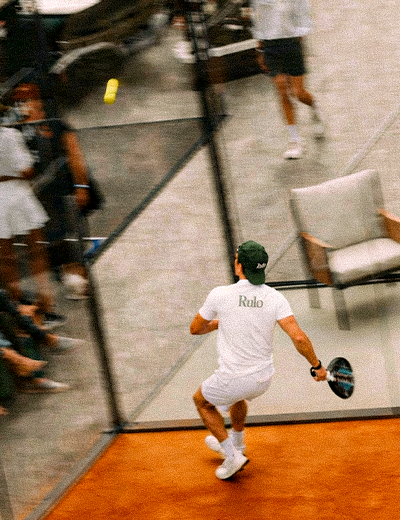 Man in white shirt and cap playing paddle tennis on orange court near net and chair.