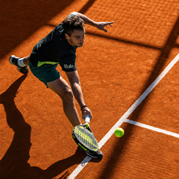 Male tennis player in black and green lunges to hit ball on a clay court.