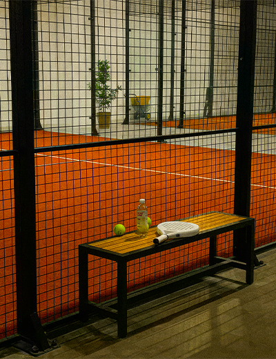 Bench with a tennis paddle, ball, and water bottle beside an indoor orange paddle tennis court.