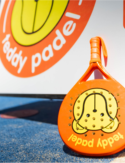 Orange Teddy Padel racket on blue padel court with matching logo in the background.