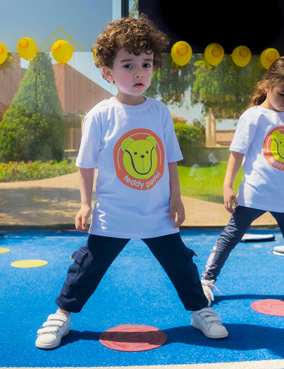 Young boy with curly hair wearing a white Teddy Patrol t-shirt standing on colorful circles outdoors.