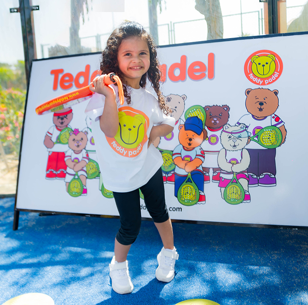 Smiling girl holding orange paddle stands on blue court in front of teddy bear paddle banner.