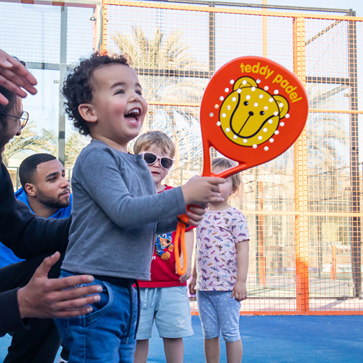 Smiling young child holding orange paddle labeled Teddy Padel while others watch on blue paddle court.