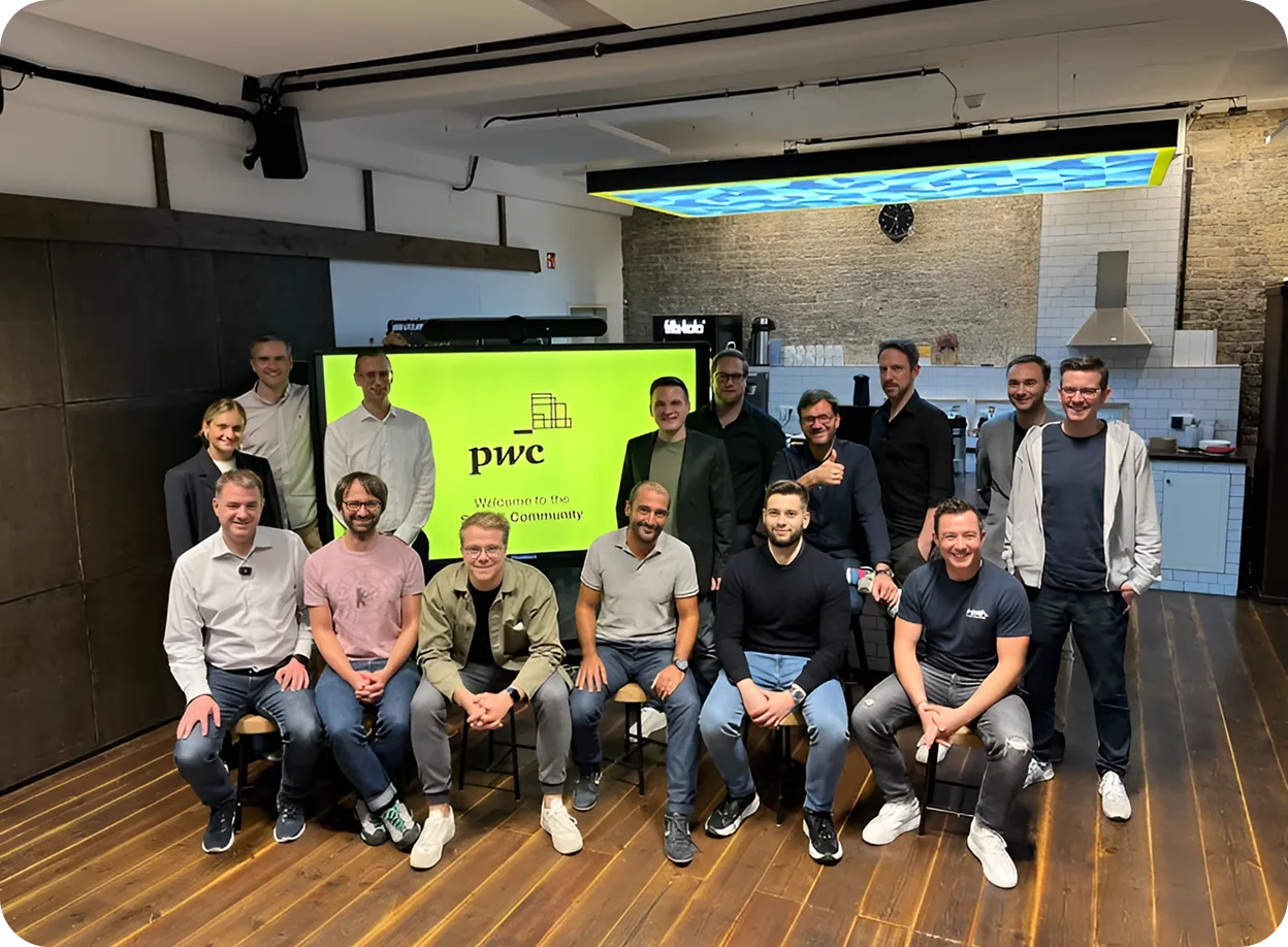 Gruppenfoto von sechzehn Personen in einem Büro mit Holzboden, einem großen Bildschirm mit dem pwc-Logo und dem Text 'Welcome to the Community'.