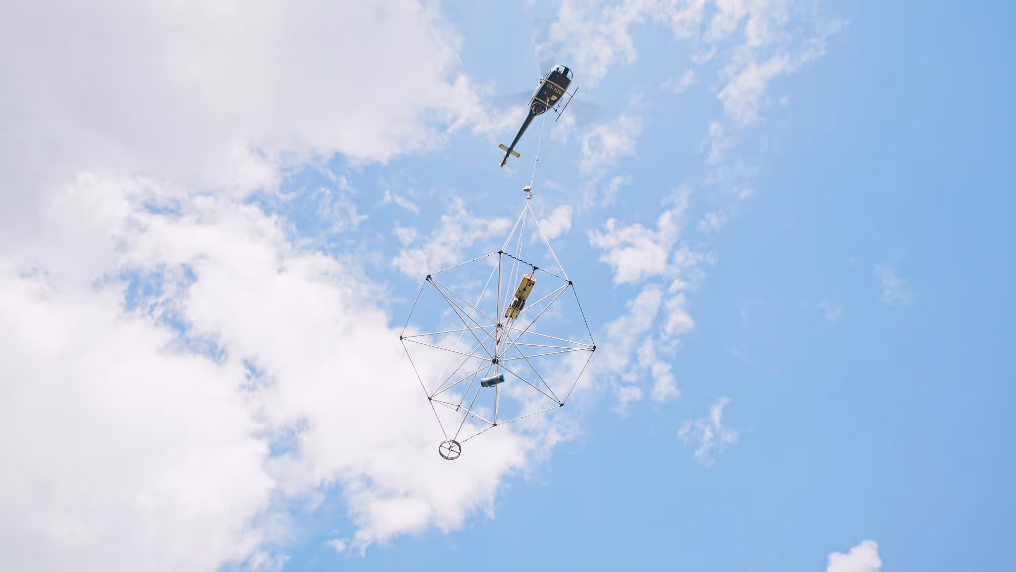 Helicopter carrying a large circular metal framework with attached equipment against a partly cloudy blue sky.