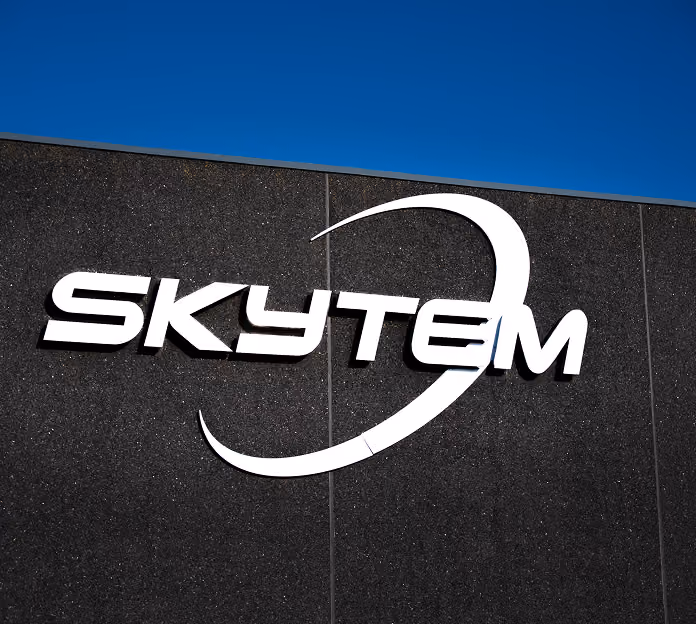 White SKYTEM logo mounted on a dark textured building wall with a clear blue sky background.