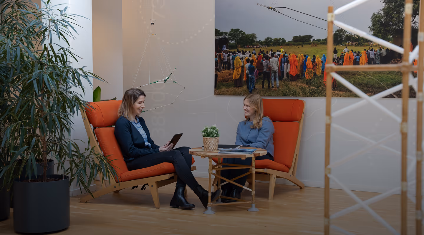 Two women sitting on orange cushioned chairs facing each other across a small round wooden table with a plant and notebooks in a modern office setting.