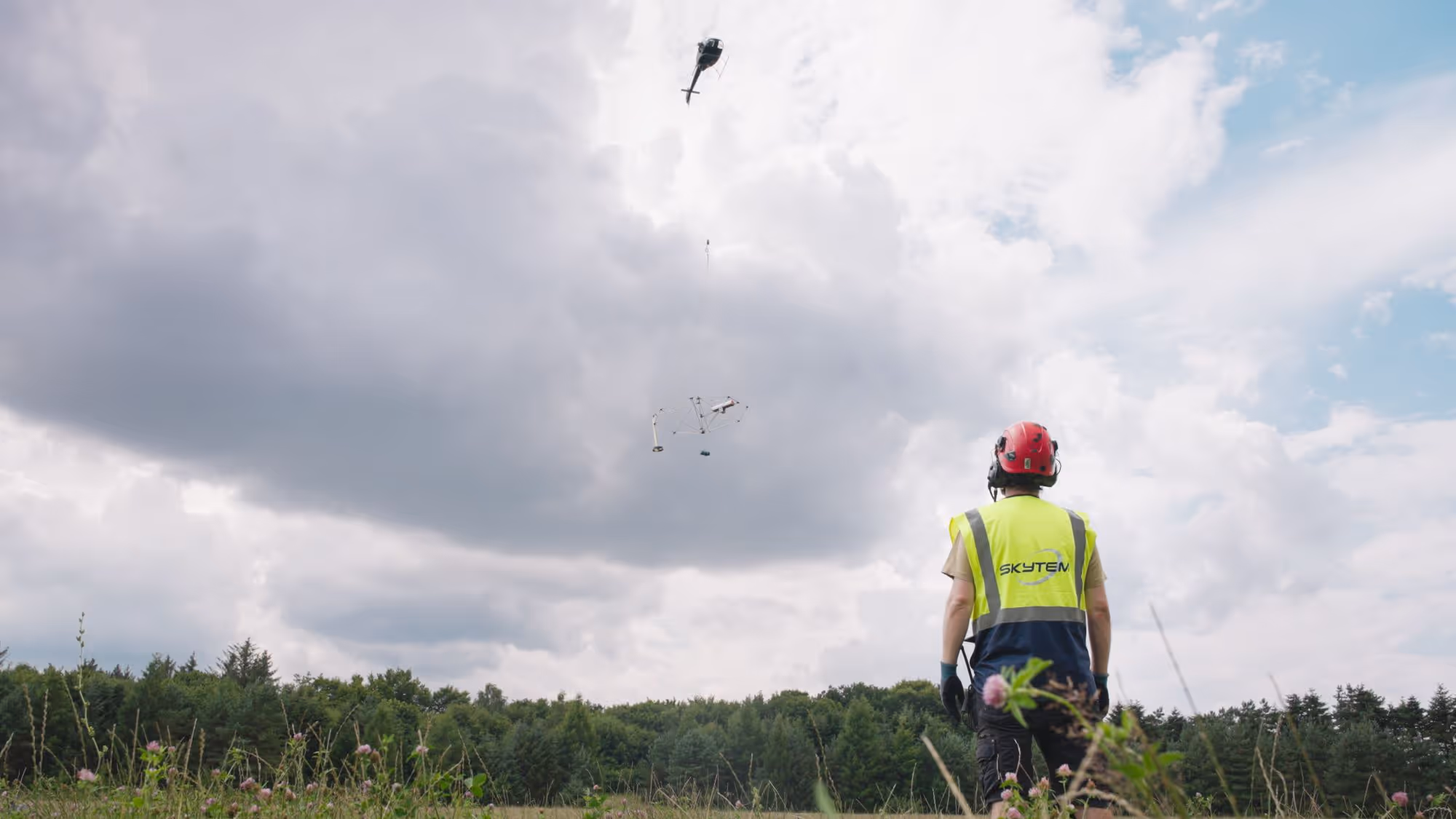 Worker in a red helmet and yellow vest supervising a helicopter lifting a drone-like device in a grassy field with trees under a cloudy sky.
