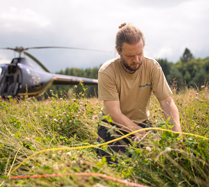 Man in a tan shirt working in a grassy field with a helicopter in the background.