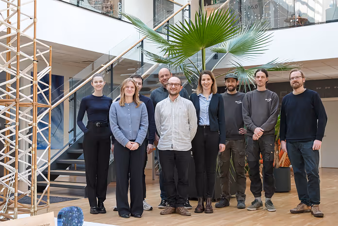 Group of nine diverse colleagues standing indoors in a modern office with plants and stairs in the background.