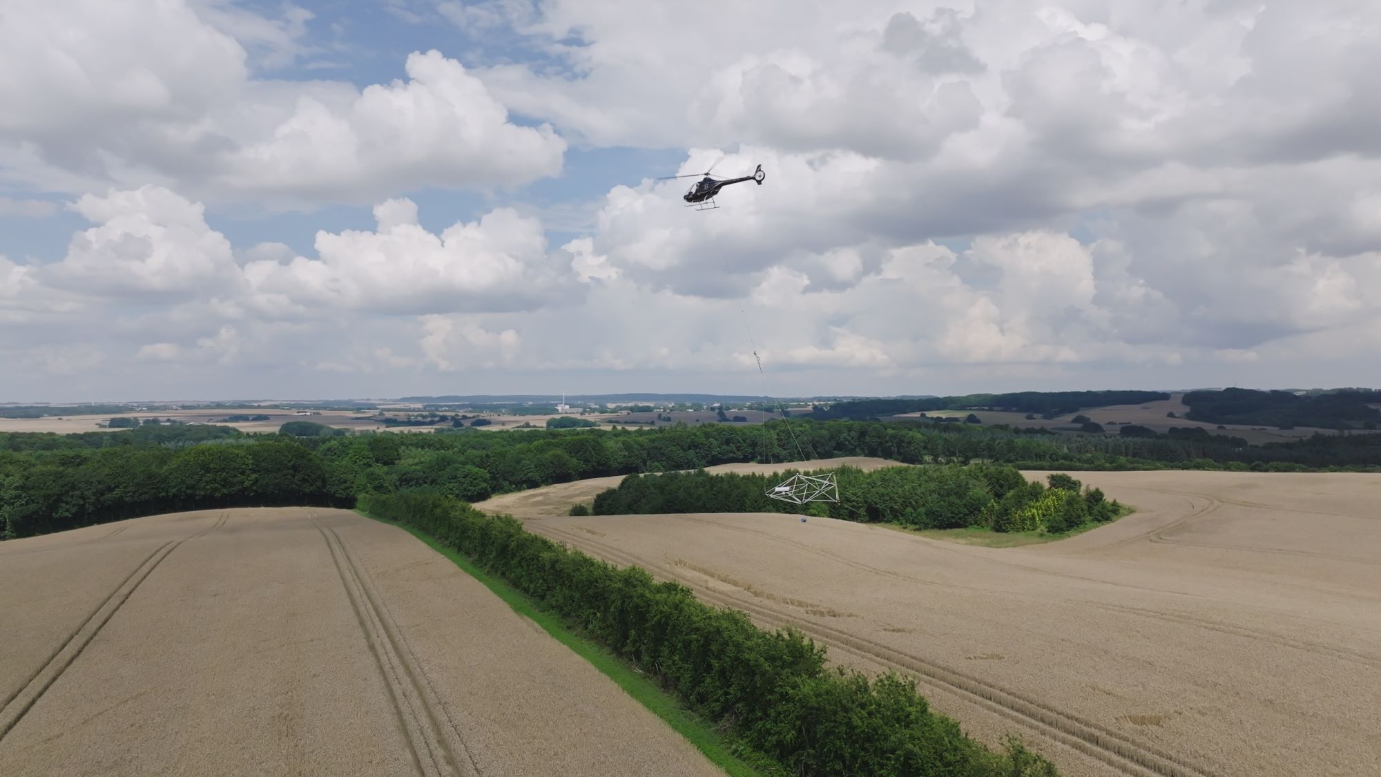 Helicopter lifting a large metal structure over expansive farmland with trees under a cloudy sky.