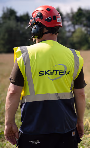 Person wearing a red safety helmet, ear protection, and a high-visibility vest with the 'SKYTEM' logo on the back, standing outdoors.