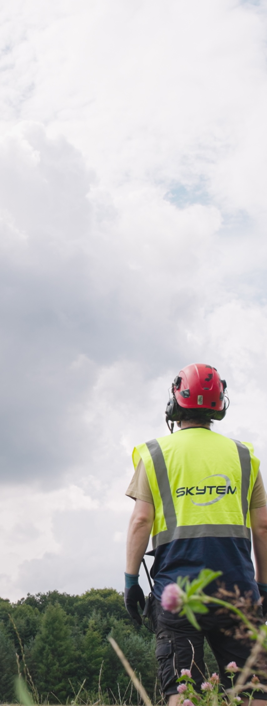 A worker wearing a red helmet and yellow vest with Skytem logo stands in a field under cloudy sky.