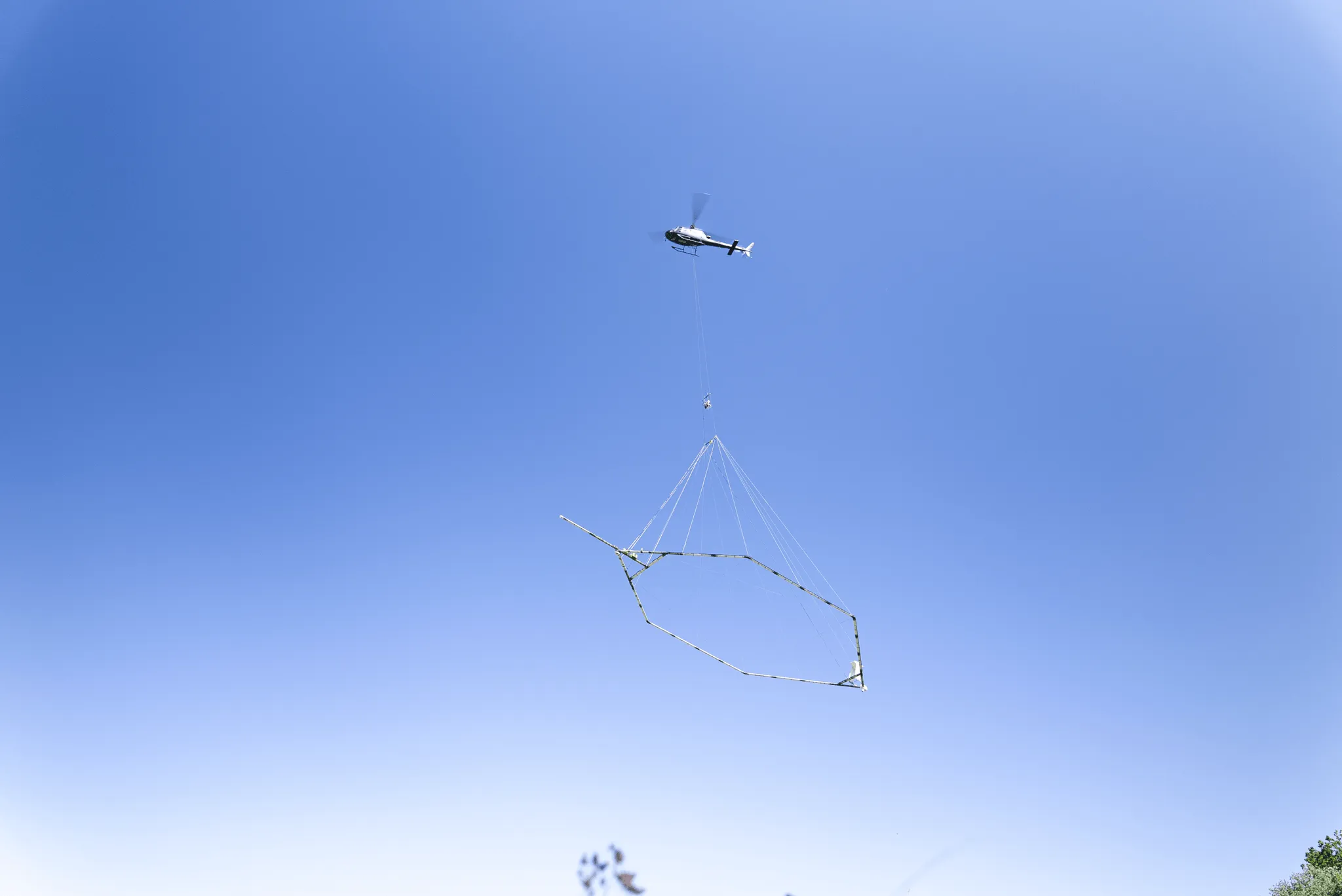 Helicopter flying in clear blue sky lifting a large geometric metal frame suspended by cables.