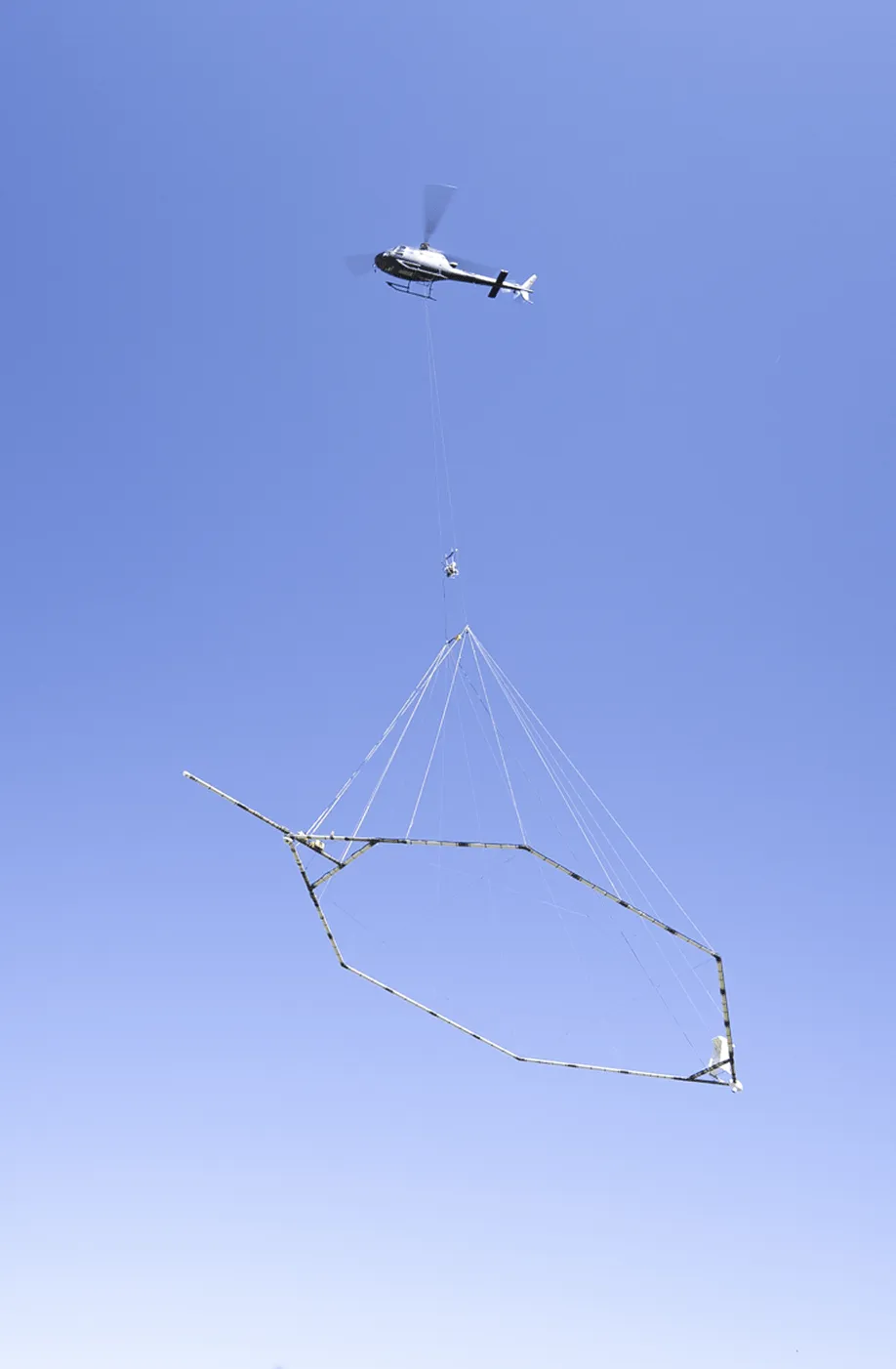 Helicopter flying with a large hexagonal frame suspended by cables against a clear blue sky.