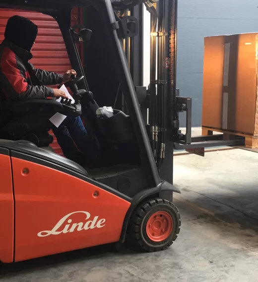 Шофьор с качулка управлява оранжев мотокар Linde, качващ голяма кутия в склад.