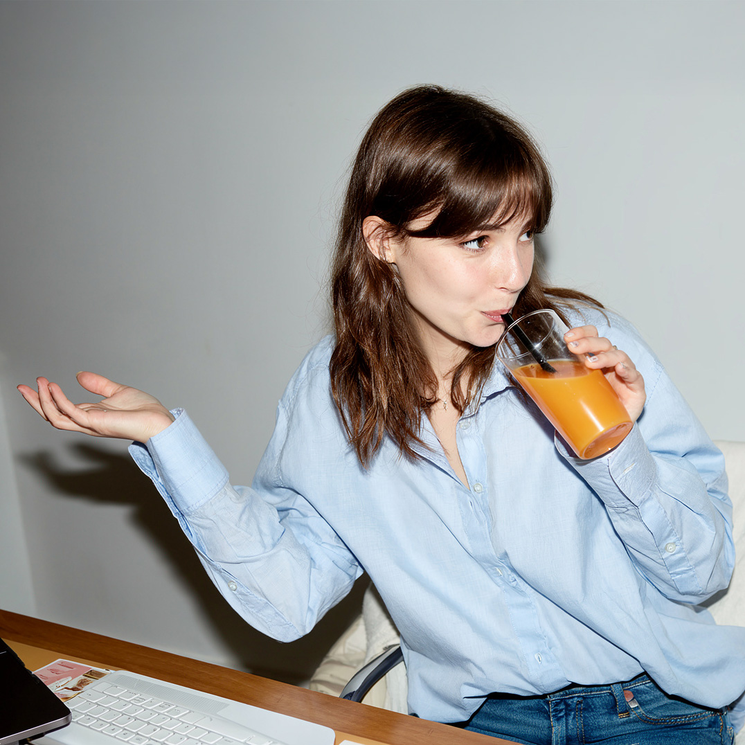 Woman in light blue shirt drinking orange juice through a straw, gesturing with her left hand while sitting at a desk with a laptop keyboard.