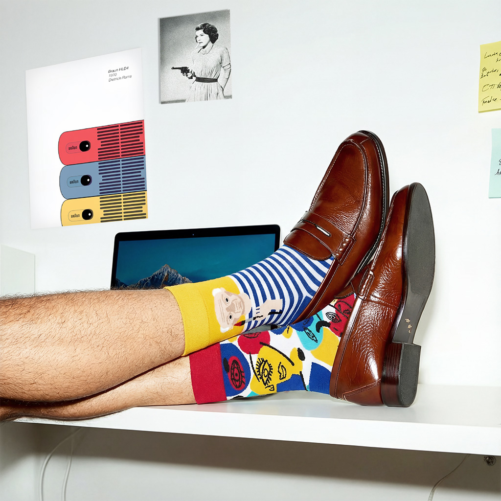 Person wearing colorful cartoon-themed socks and brown leather shoes with feet resting on a desk beside a laptop and color swatches.