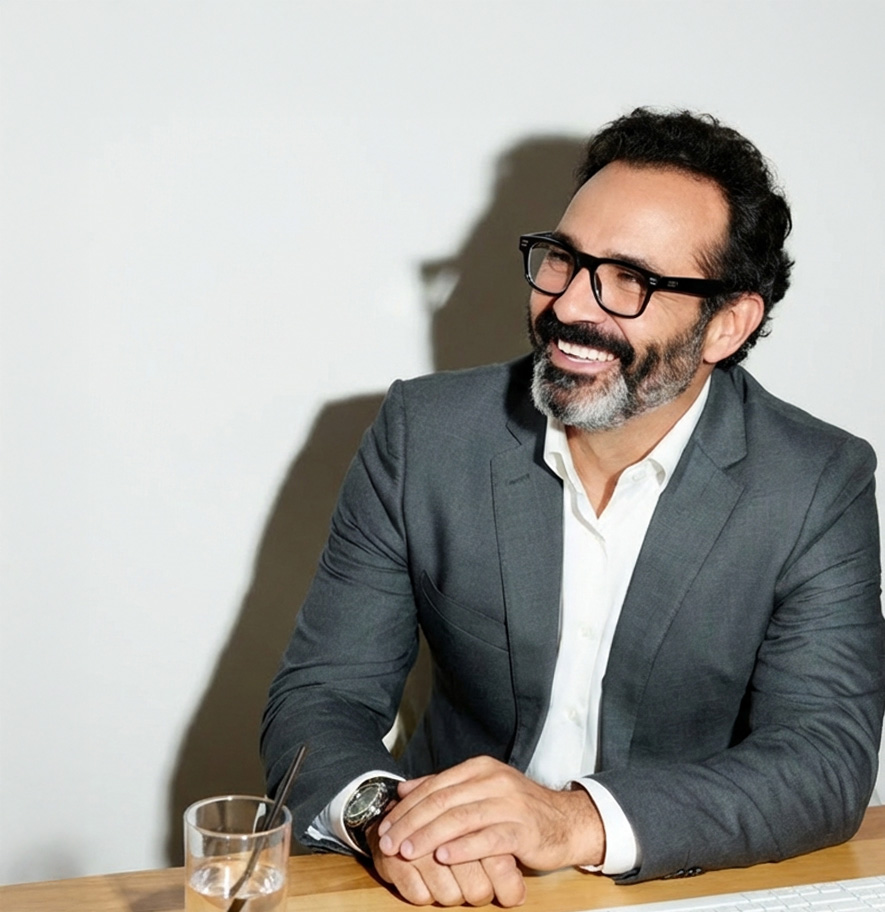 Man with glasses and beard smiling while seated at a table, wearing a dark blazer and white shirt.