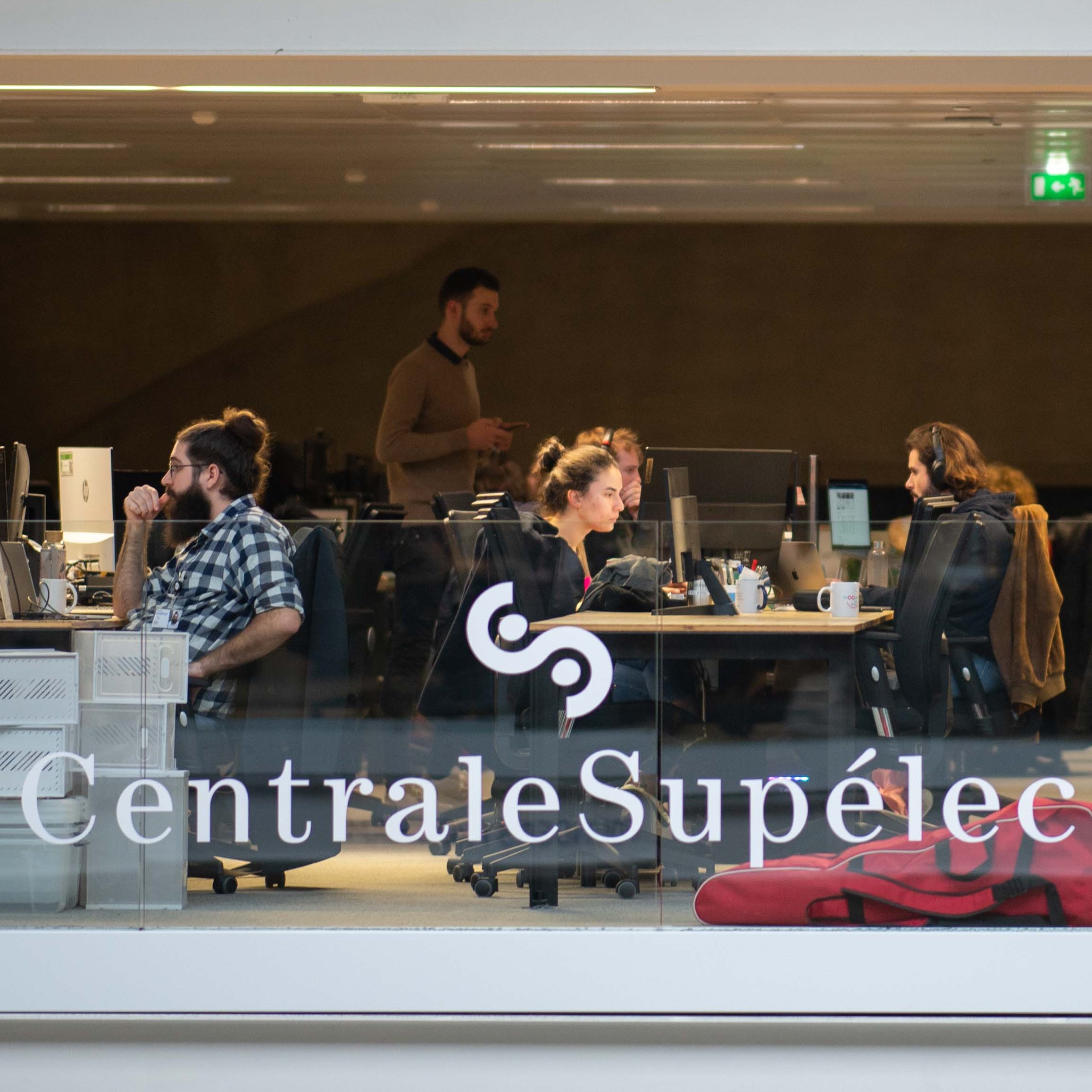 A group of people are working in a modern office with a large window displaying the company's name, surrounded by desks, computers, and a man in a black shirt.