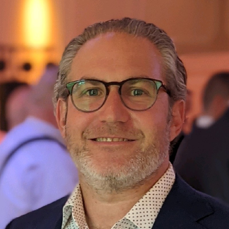 Smiling middle-aged man with gray hair and beard wearing glasses, a patterned shirt, and a dark jacket.