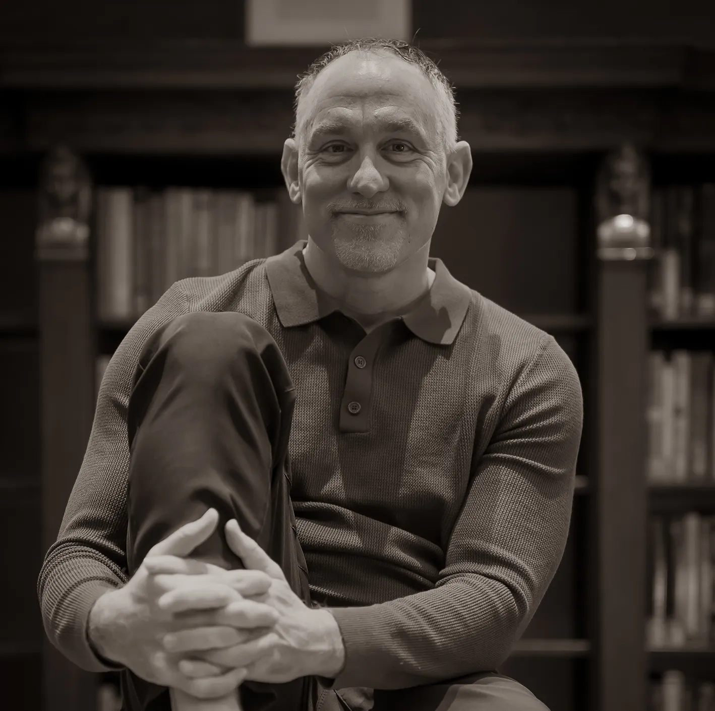 Smiling middle-aged man with short hair sitting cross-legged in front of a bookshelf.
