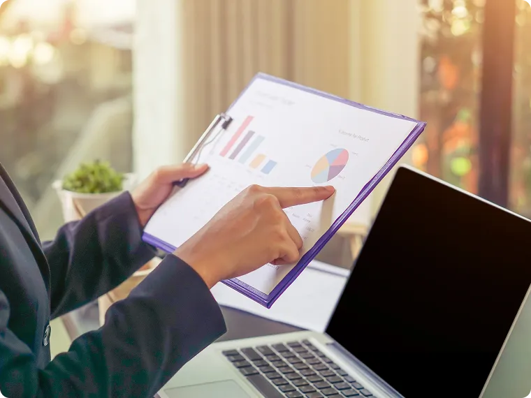Person pointing at a pie chart on a clipboard with bar charts visible, sitting near an open laptop in a sunlit room.