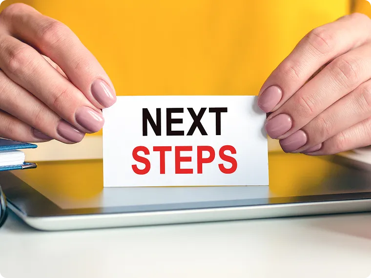 Hands holding a white card displaying the words 'NEXT STEPS' with a yellow background and a tablet underneath.