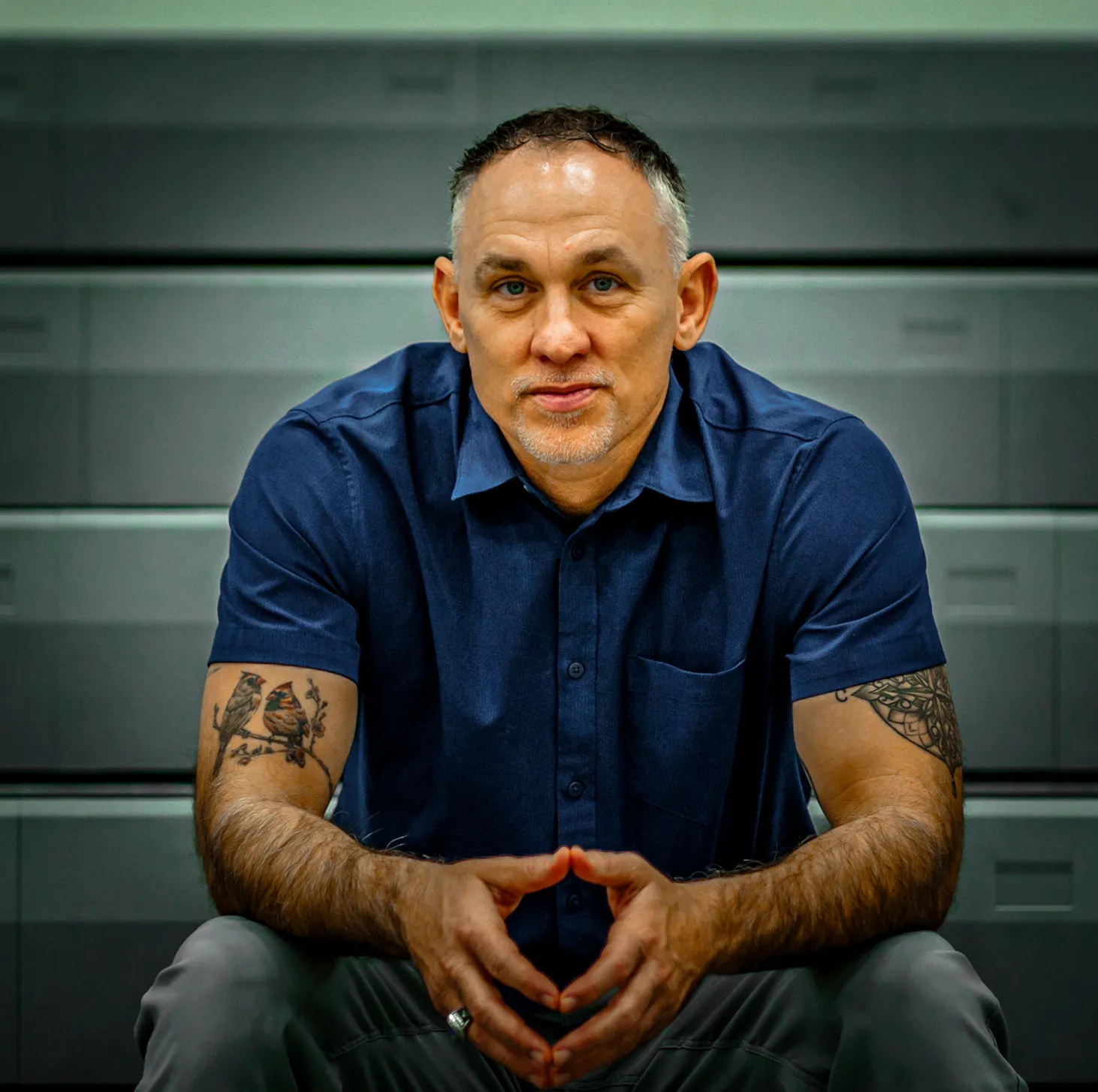 Man with short hair and tattoos on both arms, wearing a navy blue short-sleeve shirt, sitting and looking forward with hands clasped.