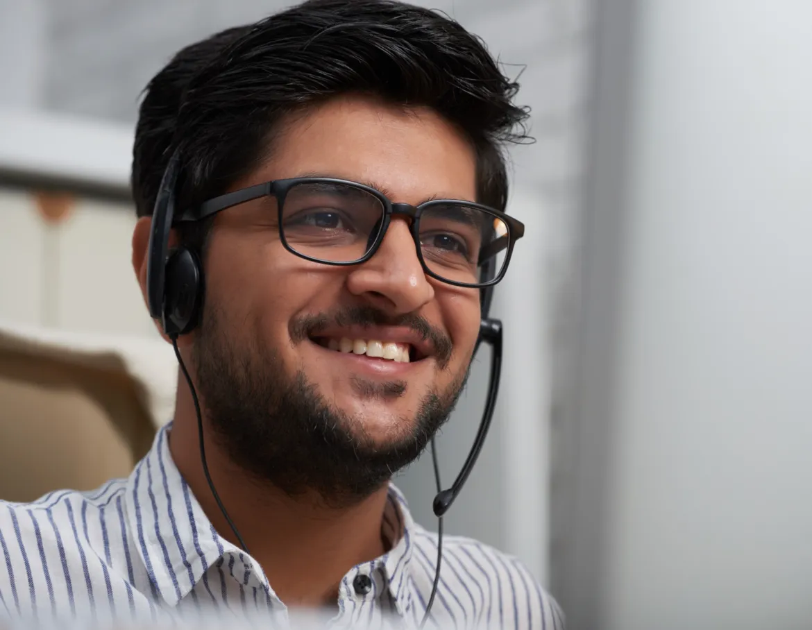 Smiling man wearing glasses and a headset with a microphone, sitting indoors.