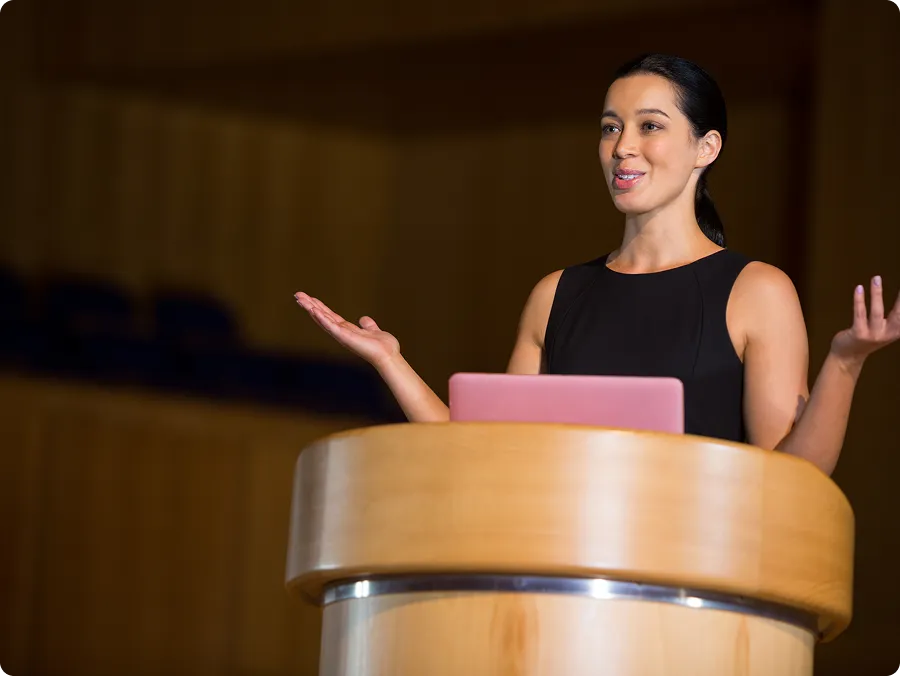 Woman in black dress speaking behind a wooden podium with a pink laptop, gesturing with open hands.