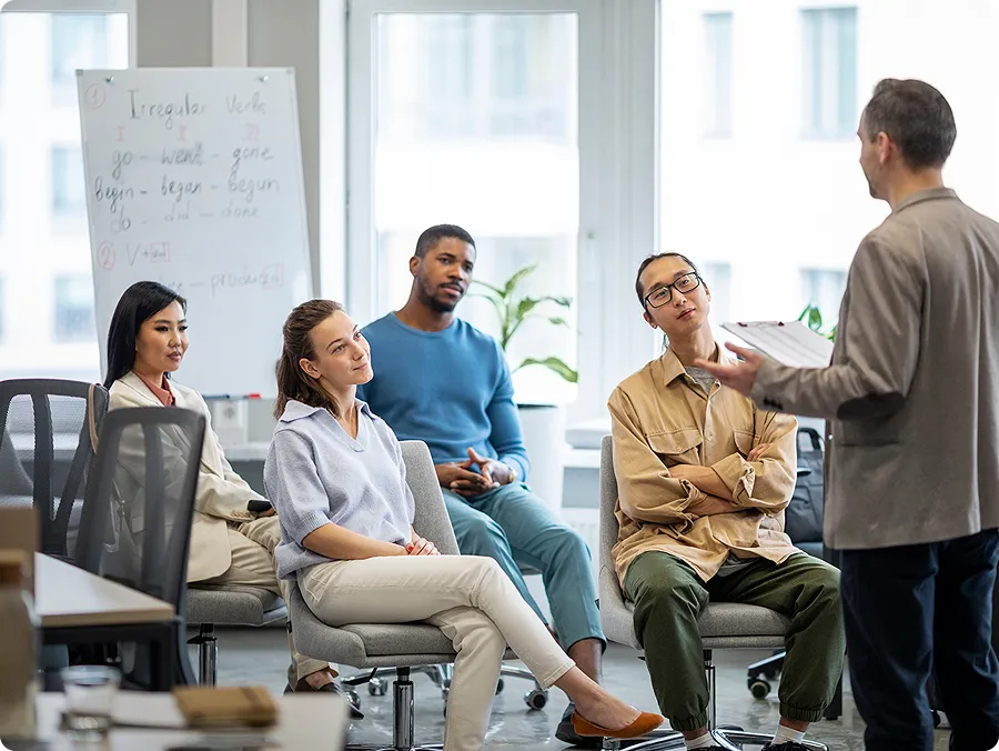 A man standing and speaking to a diverse group of four adults seated in an office setting with a whiteboard in the background.