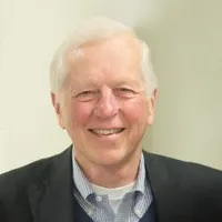 Smiling elderly man with white hair wearing a blue collared shirt and dark jacket against a light background.