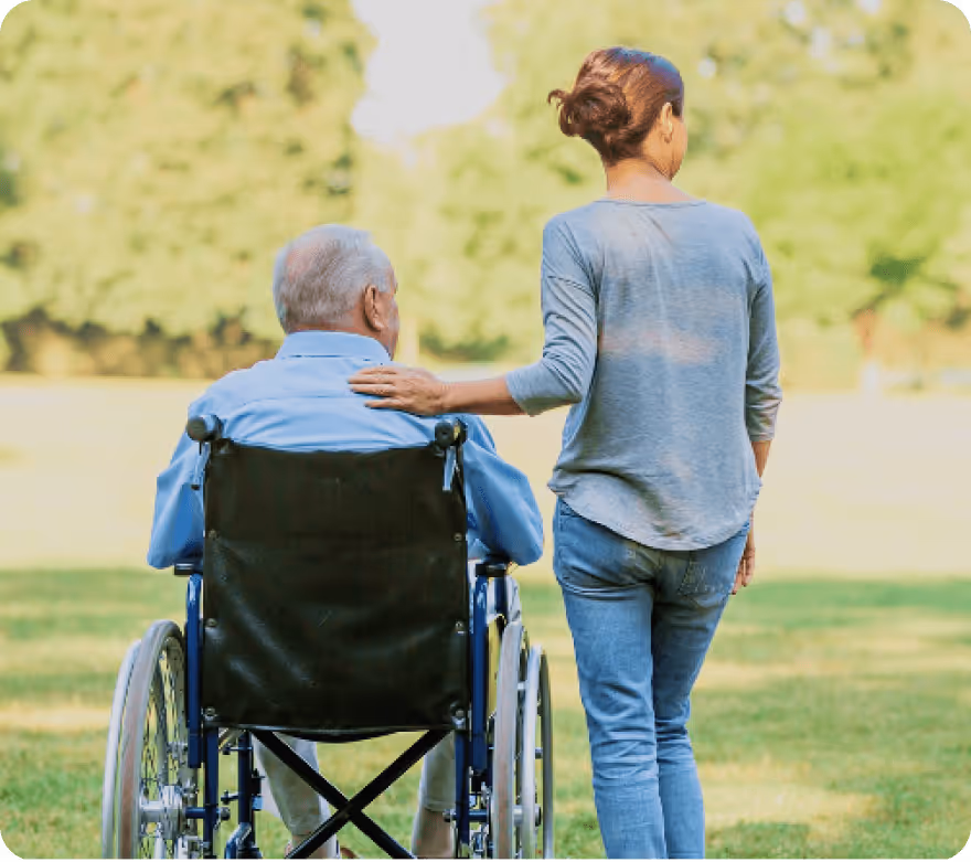 Une femme marche à côté d'un homme âgé en fauteuil roulant dans un parc, sa main posée sur son épaule.