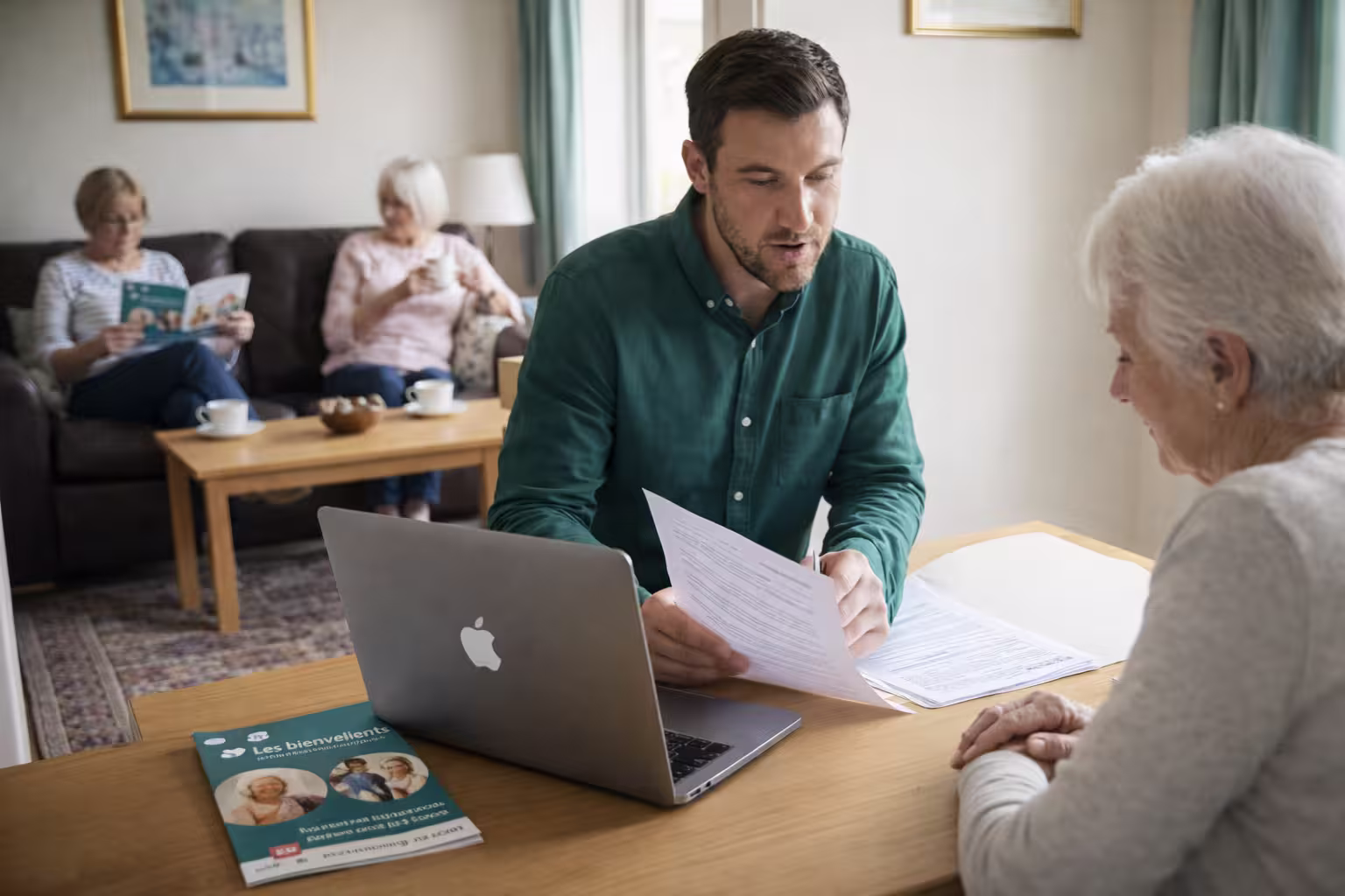 Un homme consulte des documents avec une femme âgée à une table, tandis que deux autres femmes âgées discutent en arrière-plan sur un canapé.