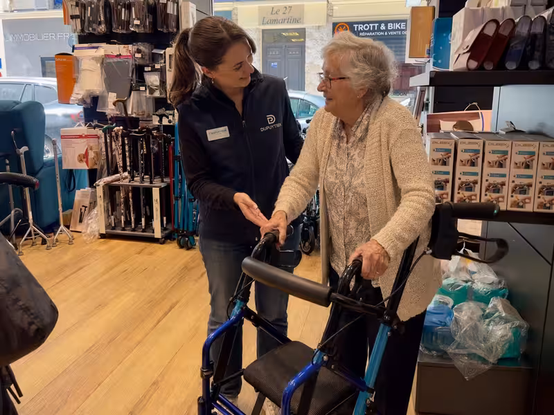 Jeune femme en uniforme Dupuytren aide une femme âgée à utiliser un déambulateur bleu dans un magasin d'aides à la mobilité.