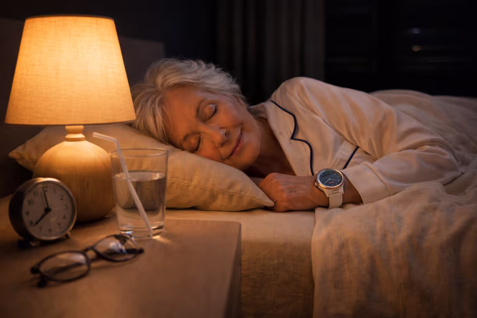 Femme âgée dormant paisiblement dans son lit la nuit avec une lampe allumée sur la table de nuit à côté d'un verre d'eau, d'une horloge et de lunettes.
