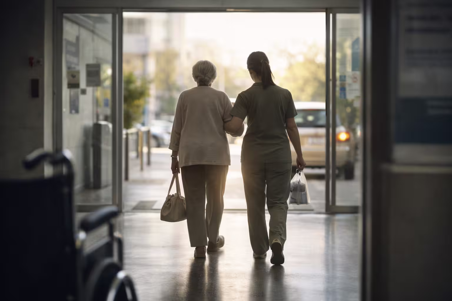 Photo d'une personne sortant de l'hôpital avec un sac, accompagnée par une auxiliaire qui l'aide à se diriger vers un véhicule ou un hall d'immeuble.